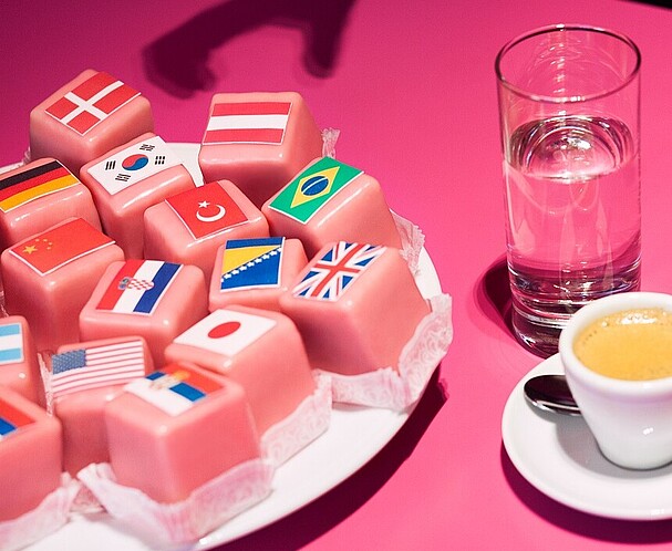 Pink pastry with flaggs beside it is a coffe and a glass of water all placed on a pink table
