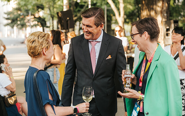 Two women and one men discussing outdoors at an event, other people in the background.