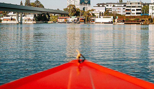 Red boat floating on river in front of it are big buildings 
